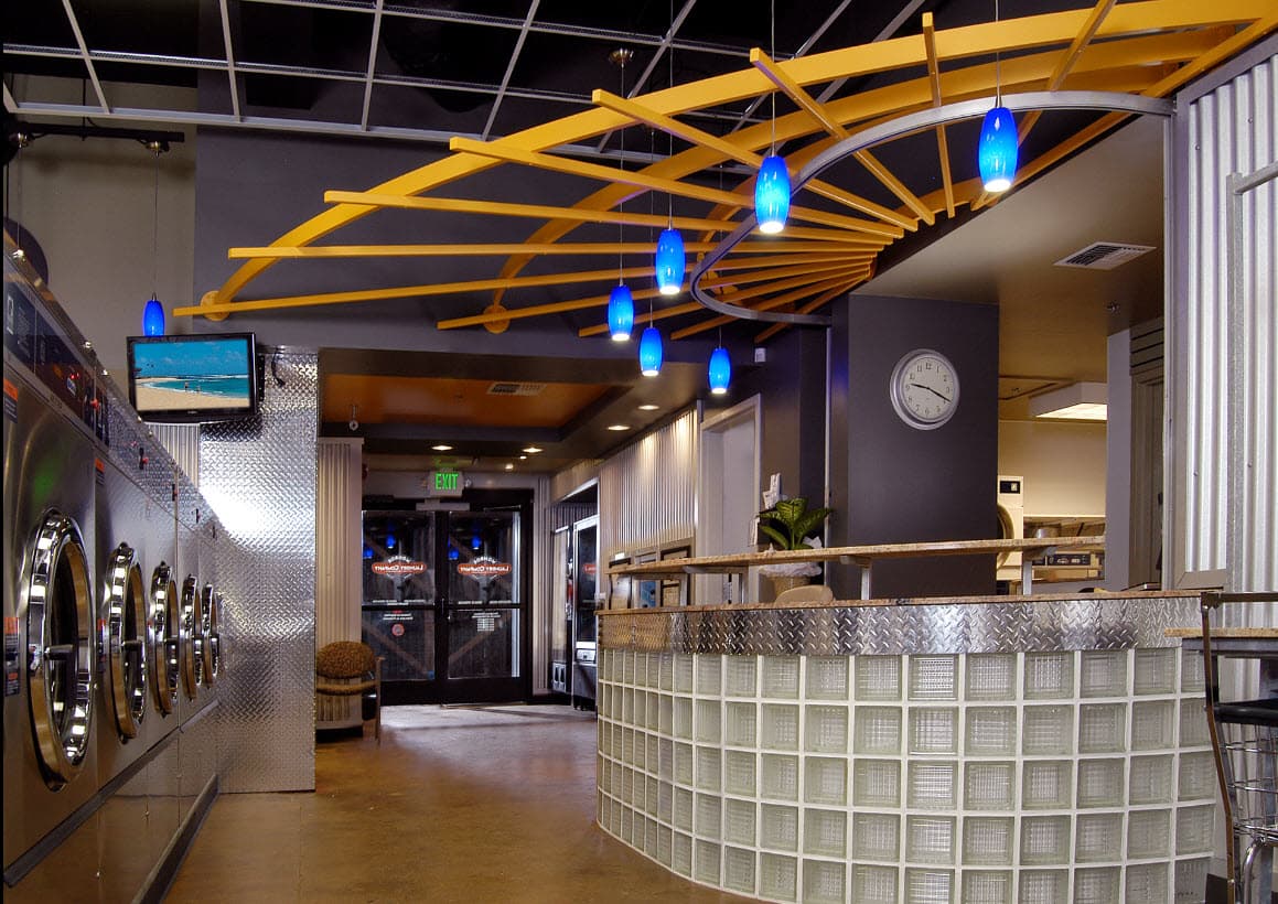 The renovated laundromat interior: a curved glass-block service counter beneath a yellow sunburst ceiling sculpture and blue pendant lights, with stainless machines along the left wall