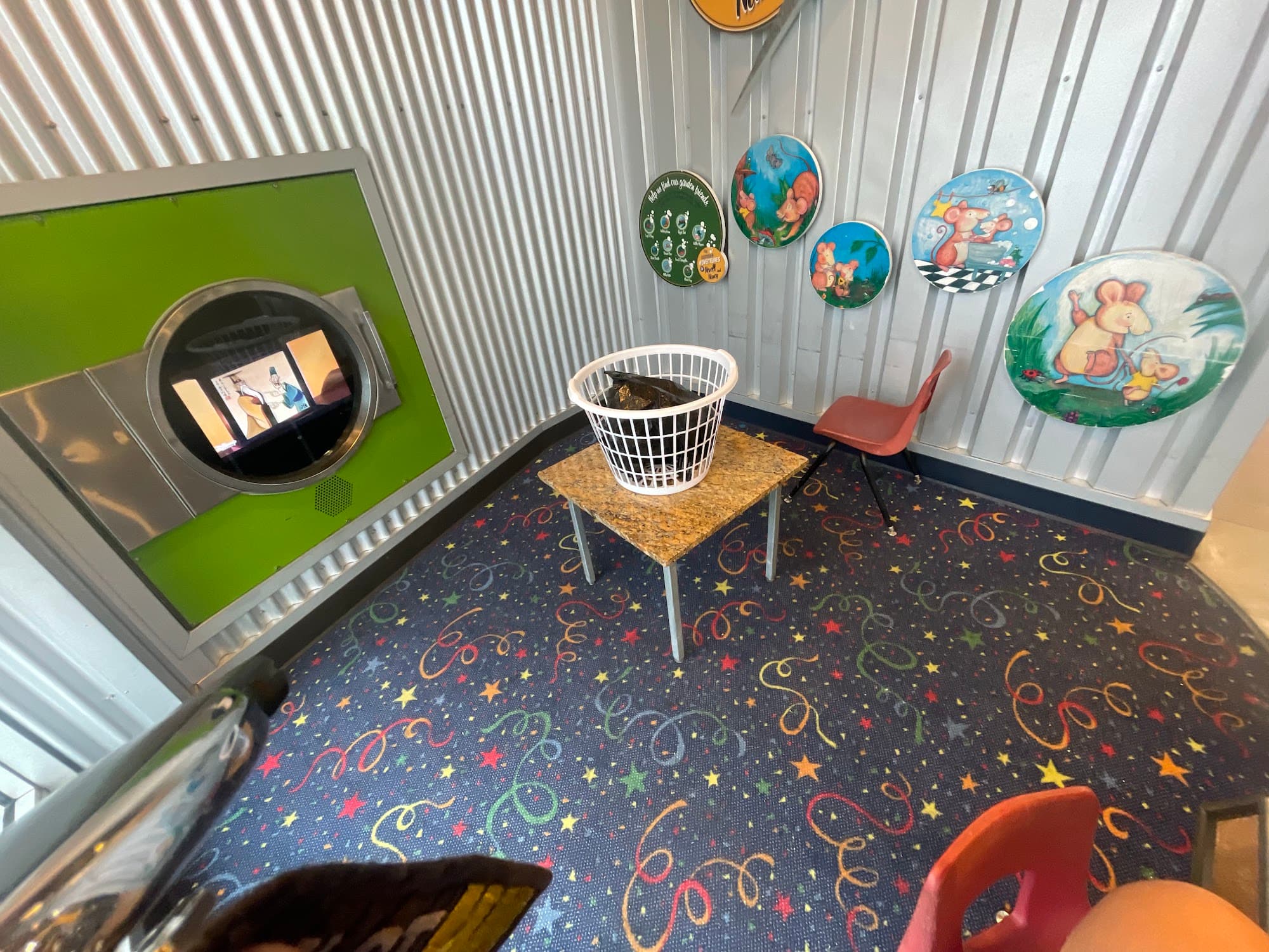 The Kids Kozy Korner: a confetti-and-stars patterned floor, a porthole-shaped TV nook in the wall, and decorative plates mounted on corrugated steel